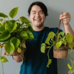 A person with a smiling face holding two pothos plants. The plant in the left hand is a large, mature pothos with trailing vines and vibrant green leaves. The plant in the right hand is a smaller, developing pothos with new growth illustration how to Grow Plants From Cuttings.